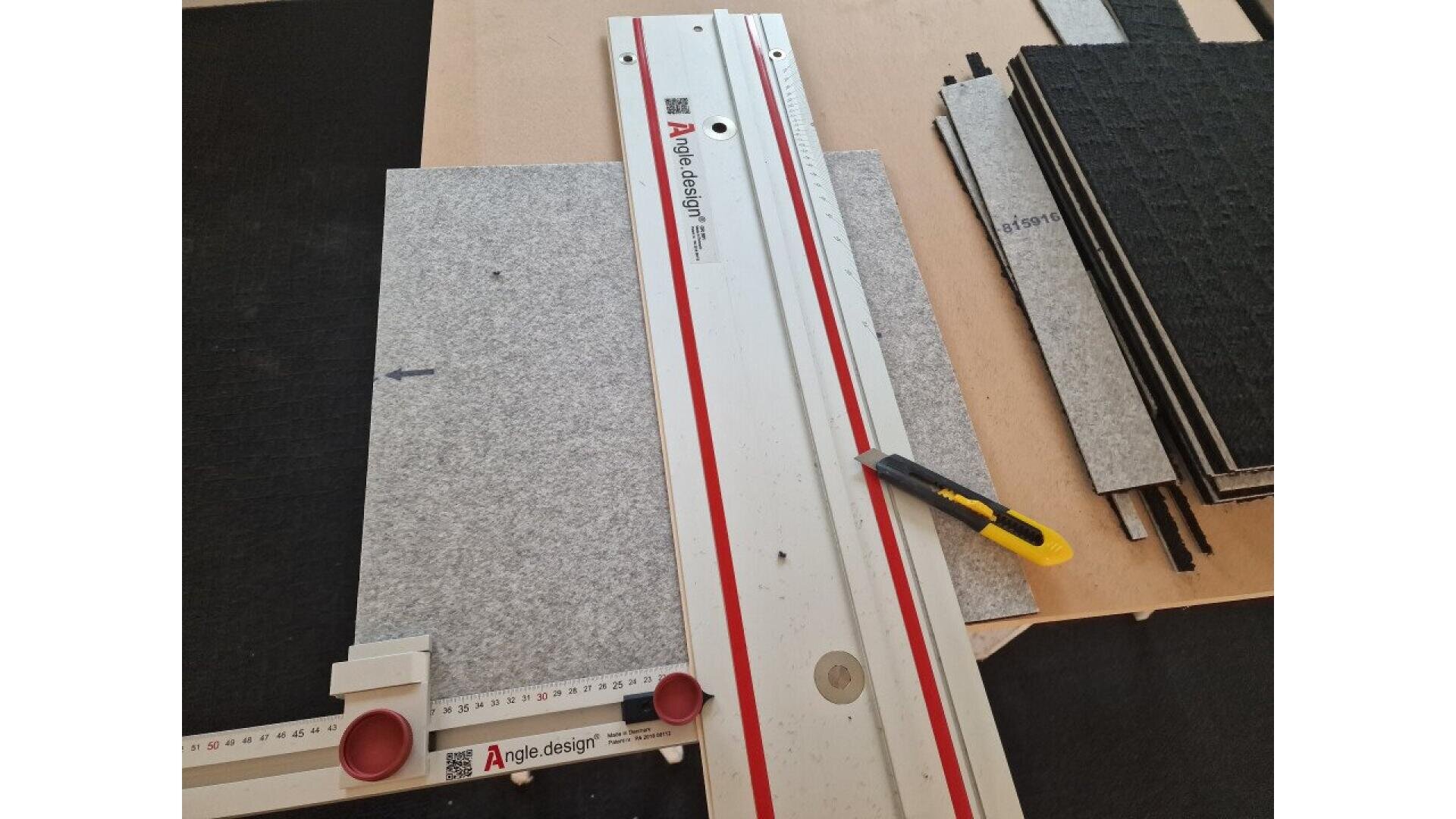 Ein Schneidwerkzeug und ein Universalmesser liegen auf einer grauen Platte und sind bereit zum Schneiden. Rechts davon befinden sich Stapel schwarzer Platten, die alle auf einer Holzoberfläche liegen. Auf der grauen Platte ist ein Pfeil eingezeichnet.