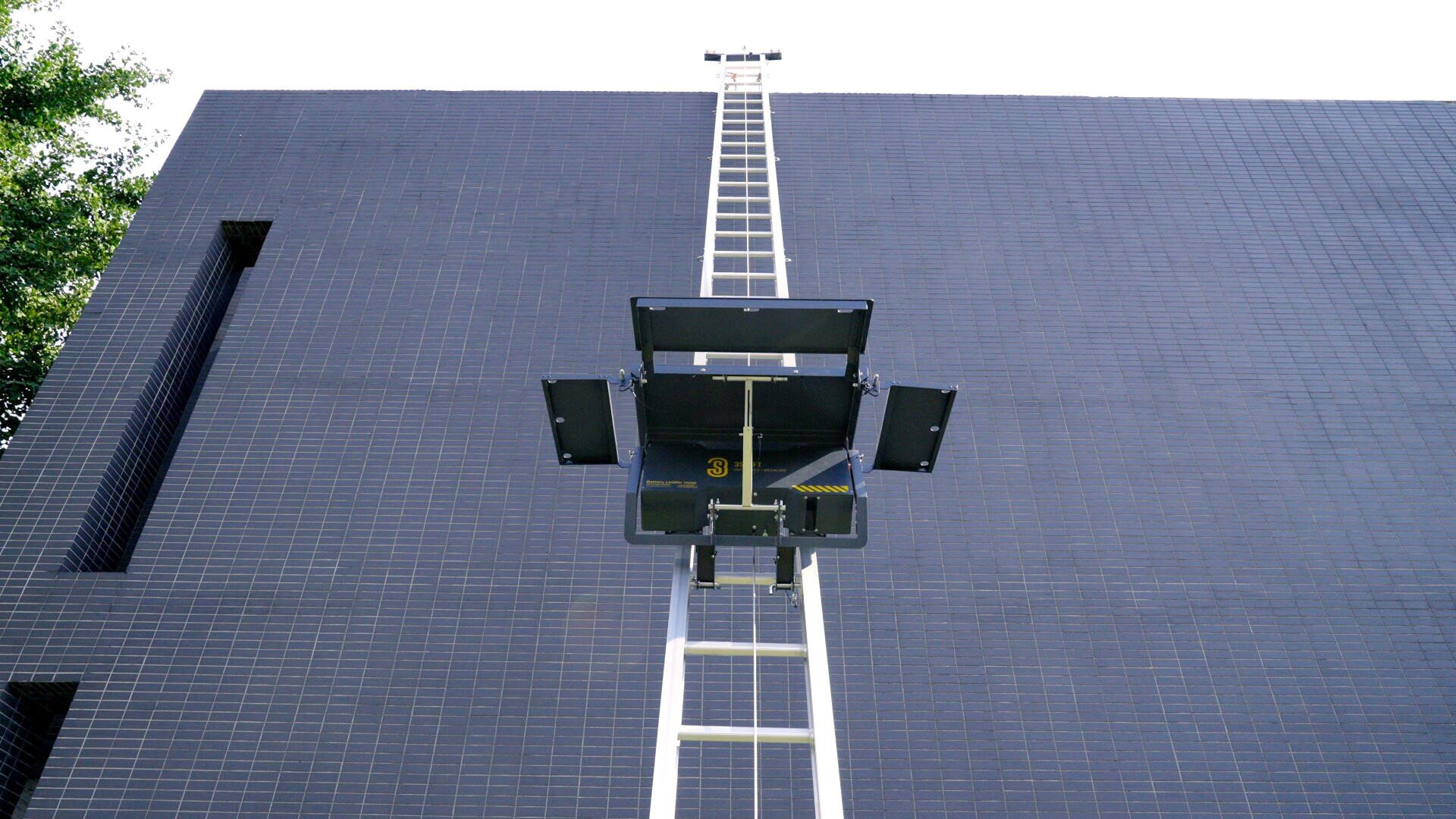 A metal ladder extends vertically up the side of a tall, dark-tiled building, ending at a small platform with open doors, seen from below against a bright sky.