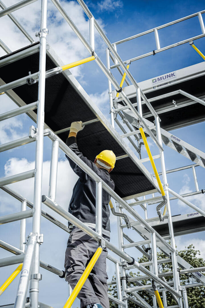 Ein Bauarbeiter mit gelbem Schutzhelm und Handschuhen hebt unter einem blauen Himmel mit Wolken eine Plattform auf eine hohe Metallgerüstkonstruktion.
