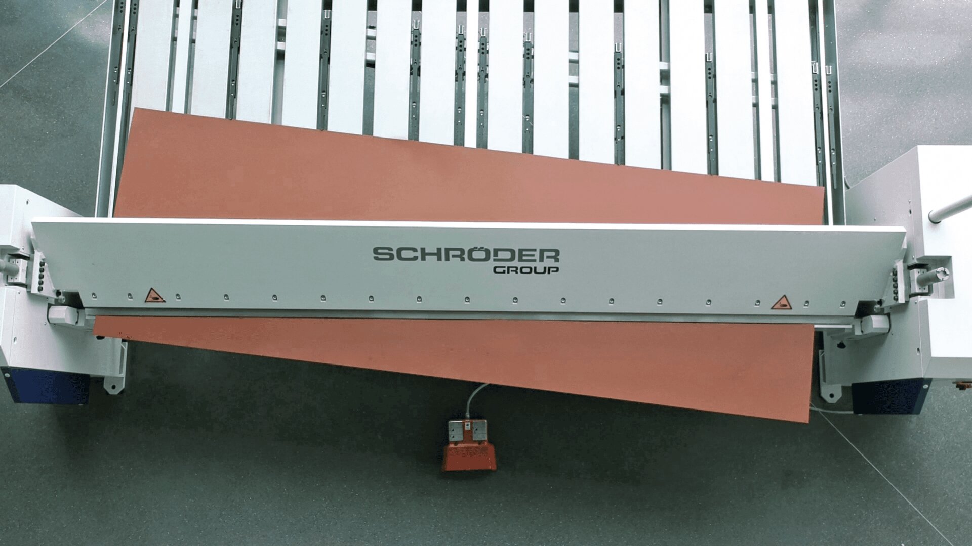 Eine Blechbiegemaschine der Schröder Group biegt ein großes rotbraunes Blech in einem Winkel, wobei unter der Maschine auf einem grauen Betonboden ein Fußpedal sichtbar ist.