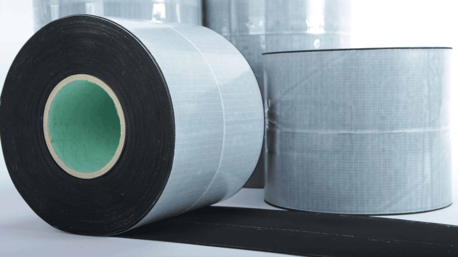 Close-up of large industrial adhesive tape rolls, with a black and gray grid-patterned surface, standing upright on a white background. One roll is lying on its side, showing a green inner core.