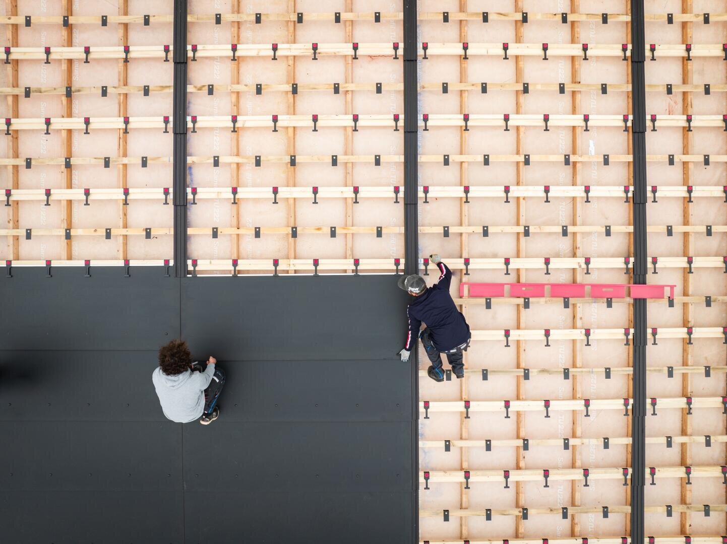 Seen from above, two people are working on a large wooden construction; one person is sitting on a black mat, the other is kneeling and reaching over the edge, surrounded by a grid of wooden planks and red staples.