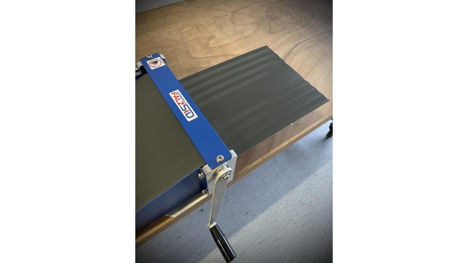 A metal sheet bending machine with a blue and silver handle labeled FACAL SCD is mounted on a wooden table, positioned over a grey floor.