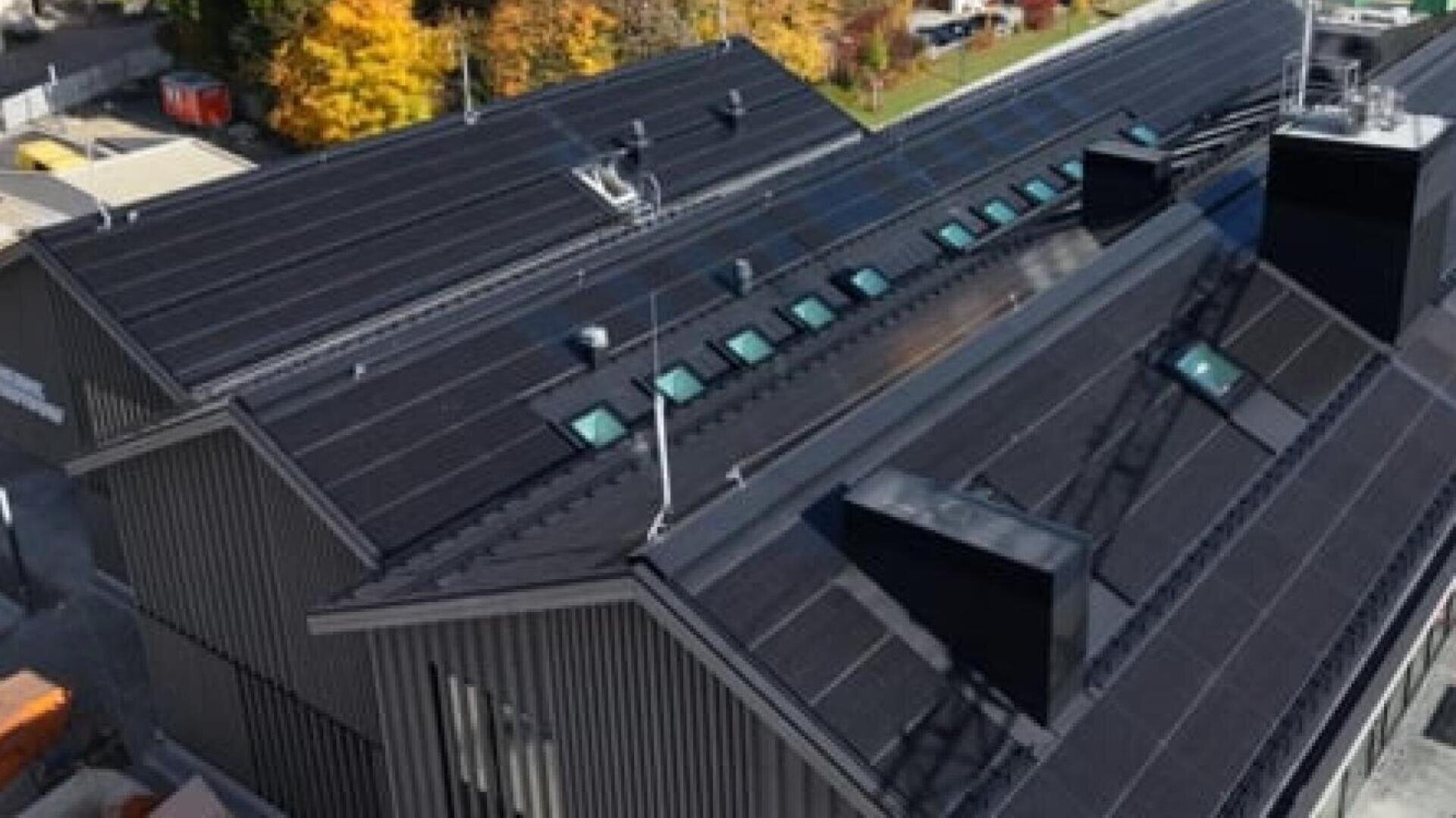 Luftaufnahme eines großen Industriegebäudes mit schwarzen Sonnenkollektoren auf dem Dach. Das Gebäude hat mehrere schräge Dachabschnitte und kleine Oberlichter; in der Nähe sind Herbstbäume und andere Strukturen zu sehen.