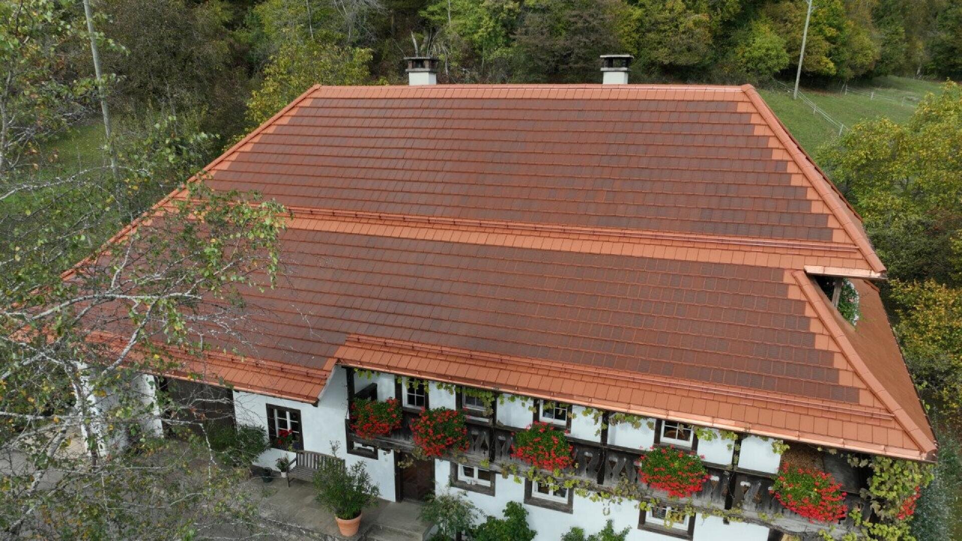 A large, traditional house with a red tiled roof and white walls, adorned with red flowers in window boxes, surrounded by green trees and foliage.