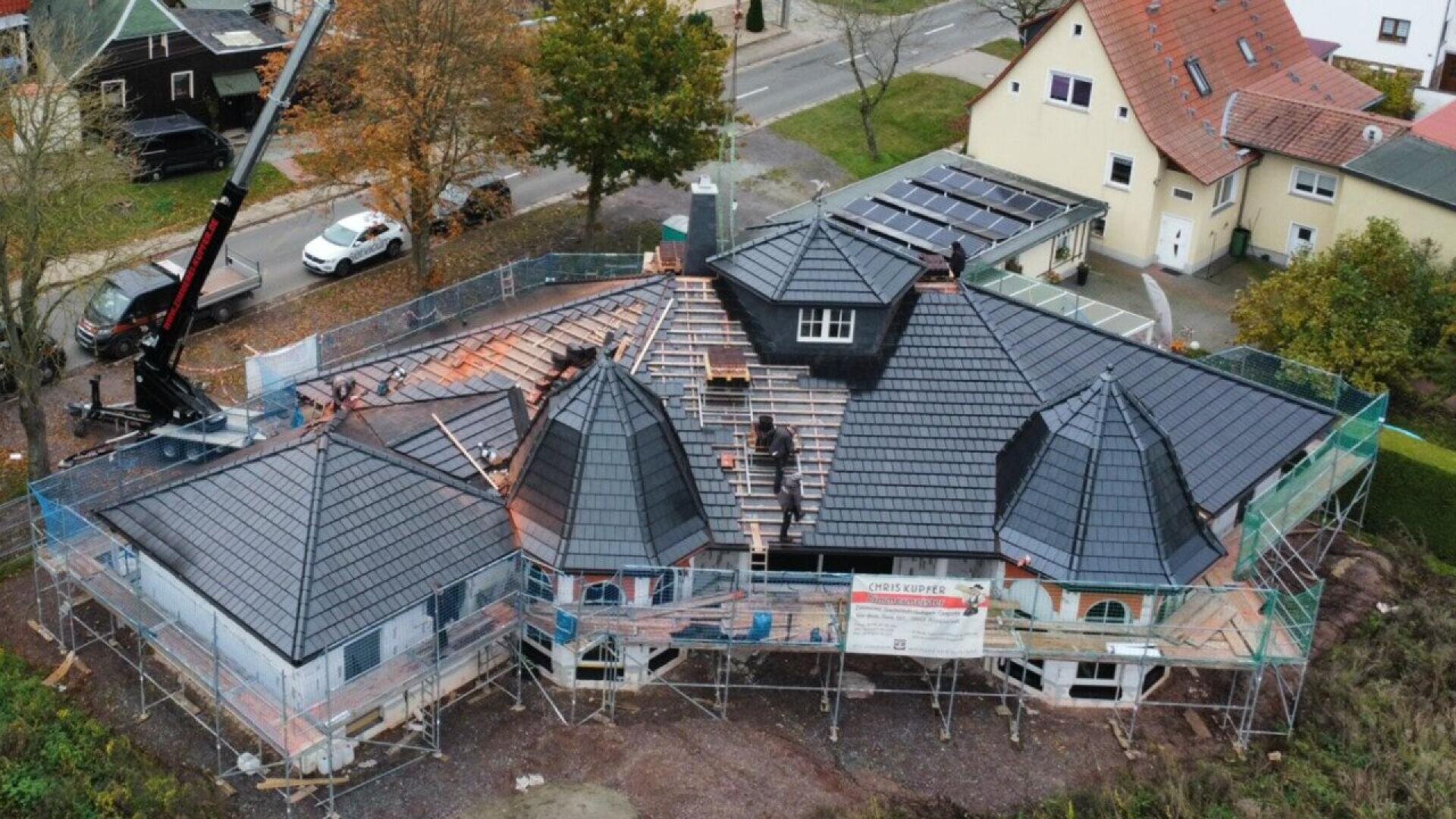 Aerial view of a large house under construction with workers installing dark roofing tiles; scaffolding surrounds the building and nearby trees display autumn colors.
