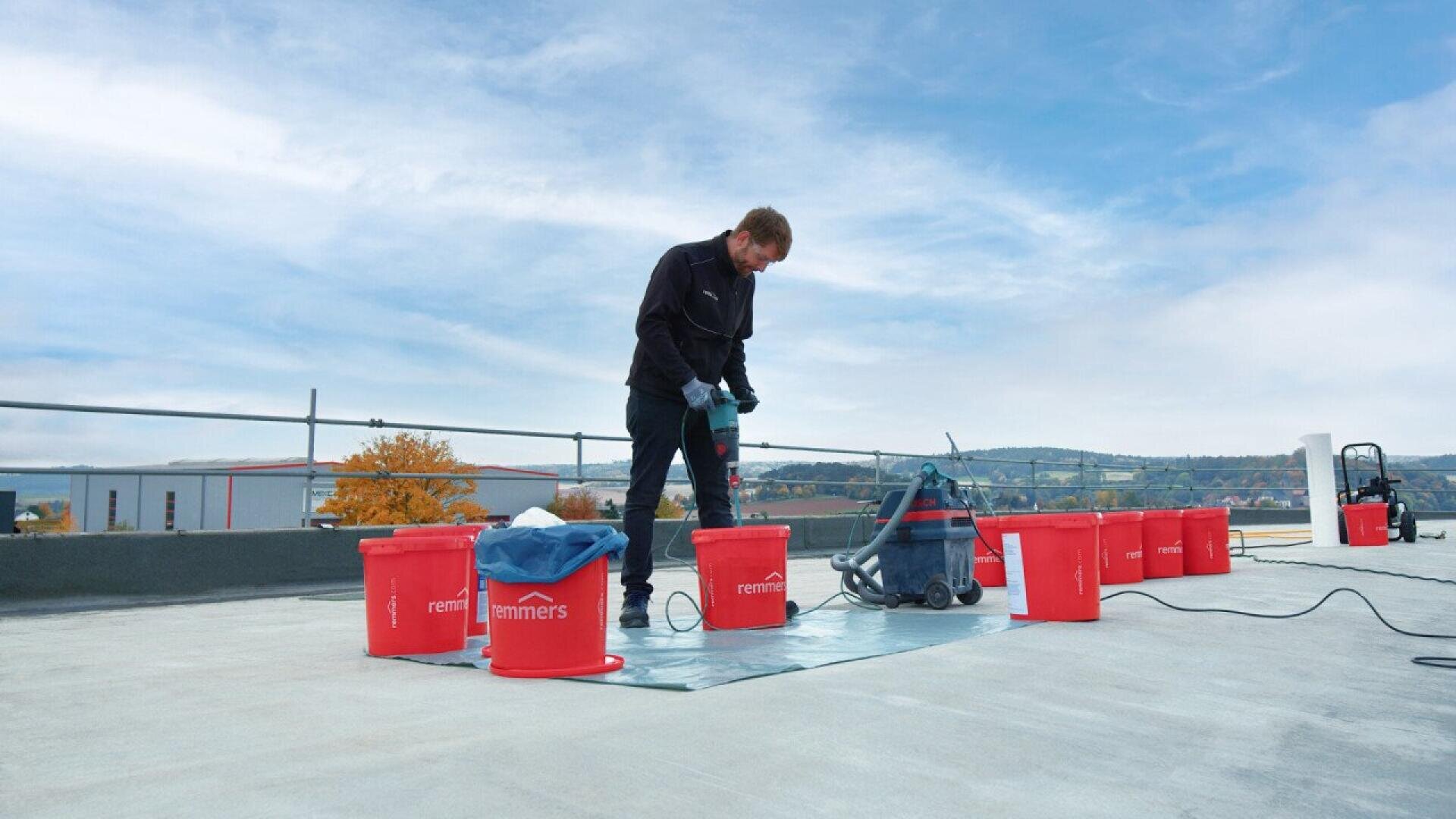 A person stands on a rooftop next to several red buckets, handling equipment for construction or maintenance work, with a blue sky and distant landscape in the background.