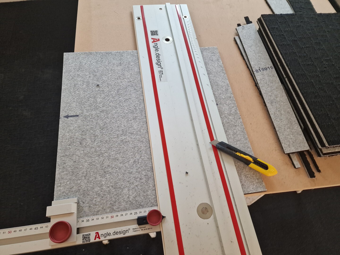 A cutting tool, a ruler guide, and a yellow utility knife are placed on a gray fabric panel, with more panels and measuring tools visible on a work surface.