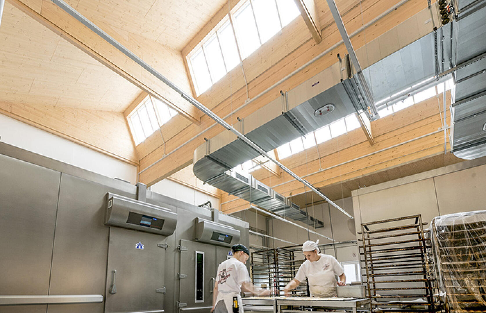 Two bakers in white uniforms work at a table in a bright, modern bakery with high ceilings, large windows, metal shelves and visible industrial equipment.
