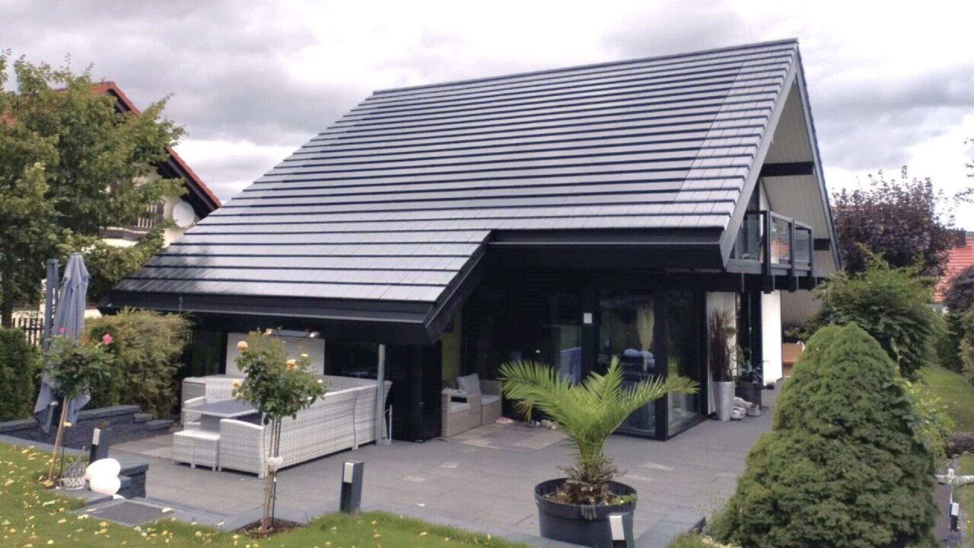A modern house with a large, sloped dark gray roof, floor-to-ceiling windows, and a patio area with white outdoor furniture. The garden features green grass, a small palm, and neatly trimmed bushes.