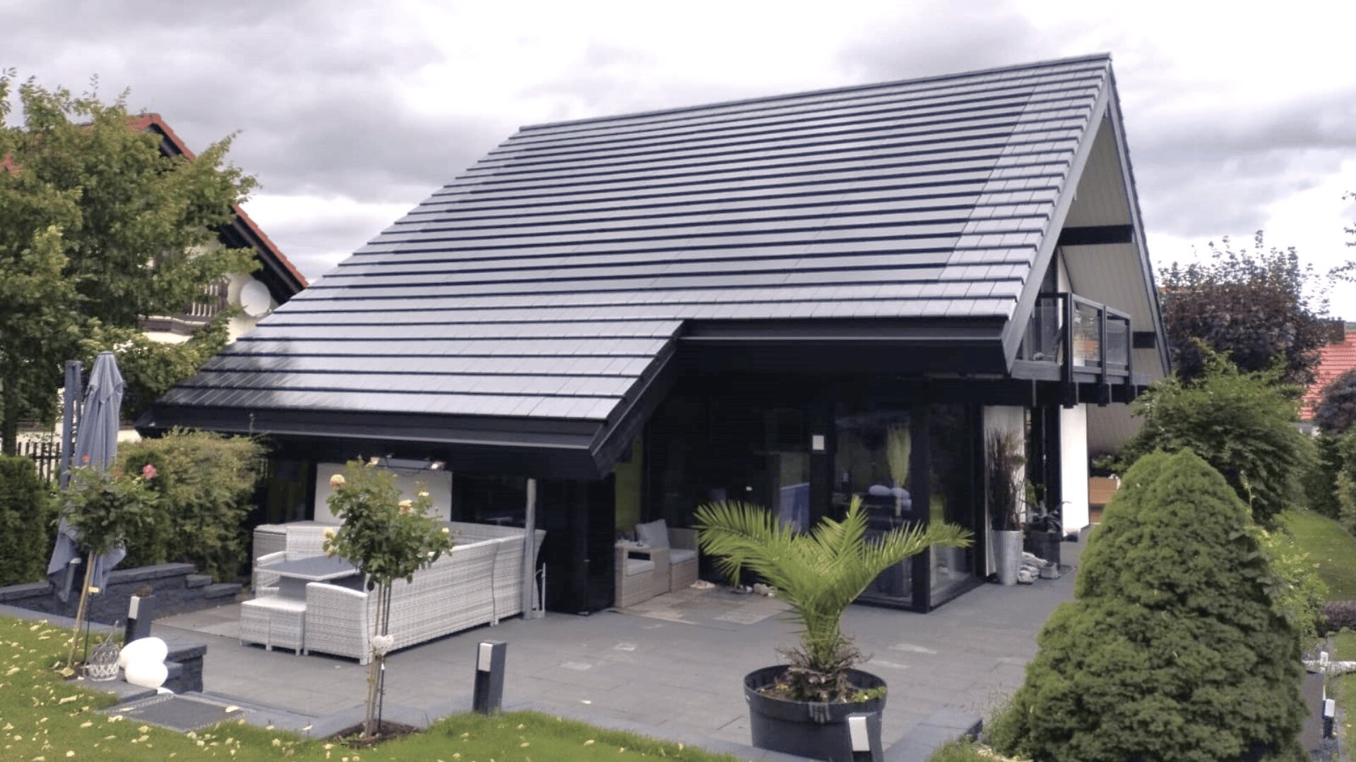 Ein modernes Haus mit einem großen Schrägdach, dunkler Außenfassade und einer Außenterrasse mit weißen Sitzgelegenheiten und Topfpflanzen, umgeben von grünem Gras und Bäumen unter einem bewölkten Himmel.