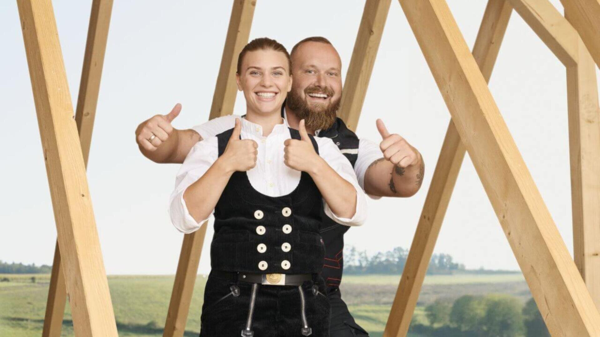 Two smiling people in traditional carpenter outfits stand outside, giving thumbs up. Wooden beams form a frame behind them, and green fields stretch into the distance under a clear sky.