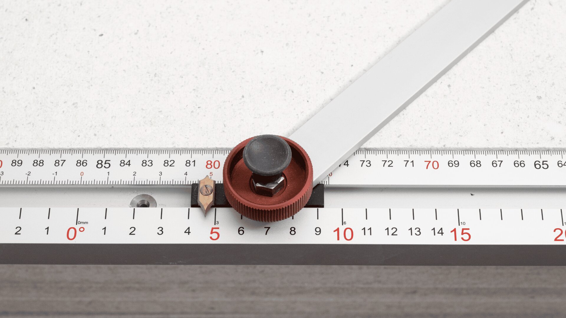 A close-up of a measuring tool with a red knob on a metal rail, marked with precise metric and degree scales, placed on a white speckled surface.