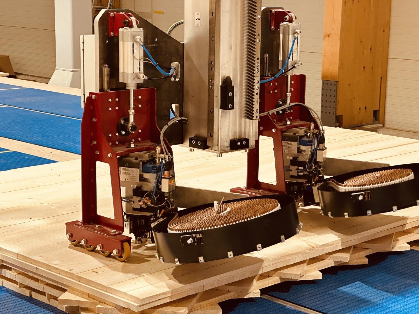 Close-up of a robot with red metal parts and cables positioned on stacked wooden planks, possibly used in construction or automated wood processing. Blue mats cover the floor in the background.
