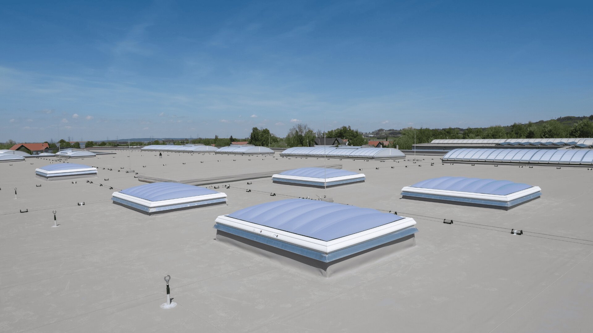 Ein flaches Industriedach mit mehreren großen, rechteckigen Oberlichtern unter einem klaren blauen Himmel; im Hintergrund sind Bäume und niedrige Gebäude zu sehen.