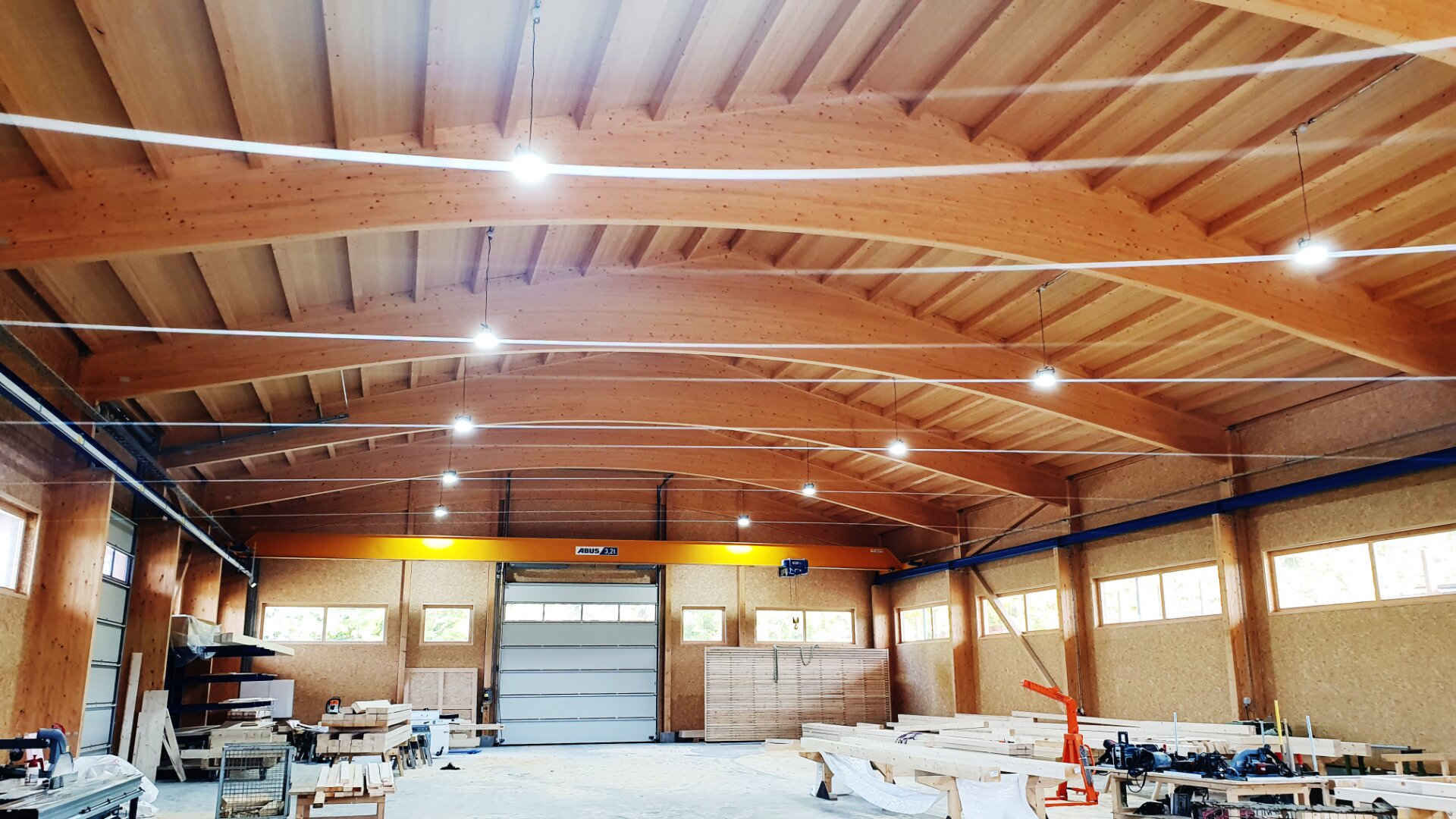 Geräumige Holzwerkstatt mit Holzbalken, hoher Decke und großen Fenstern. Verschiedene Holzbearbeitungswerkzeuge, Bretter und Bänke sind im gut beleuchteten, übersichtlichen Arbeitsbereich verteilt.