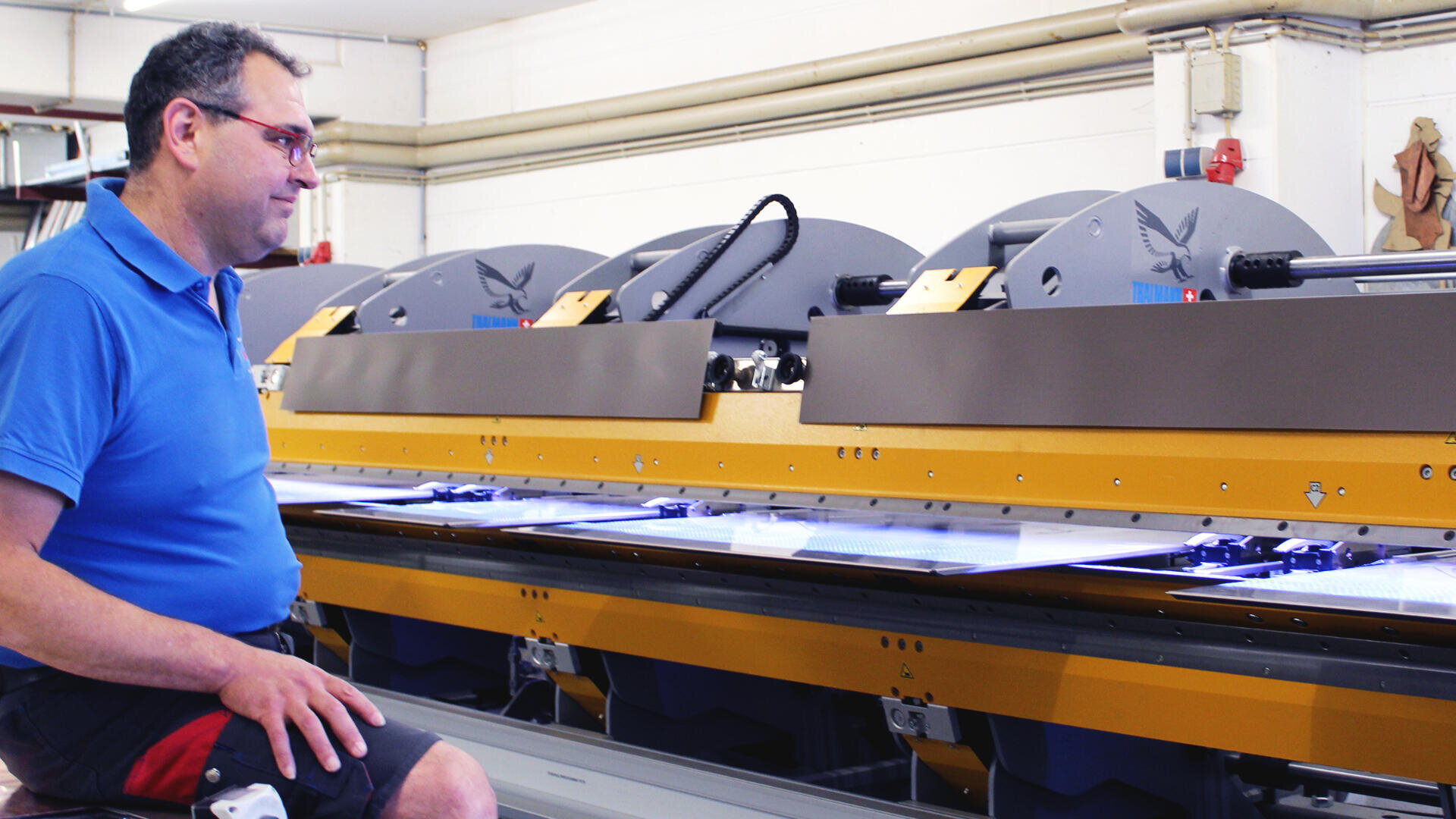 A man in a blue polo shirt and glasses sits next to a large industrial printing or cutting machine inside a well-lit factory or workshop.