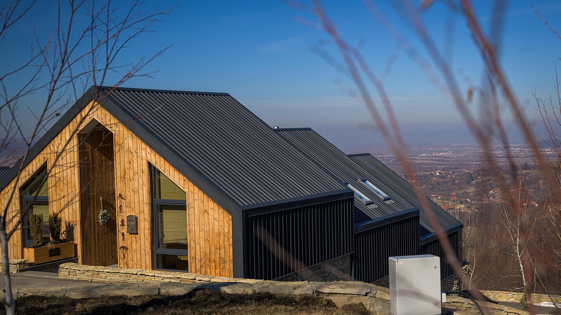 A modern wooden hut with a black metal roof stands on a hill overlooking a valley and a distant town under a clear blue sky, with bare tree branches in the foreground.