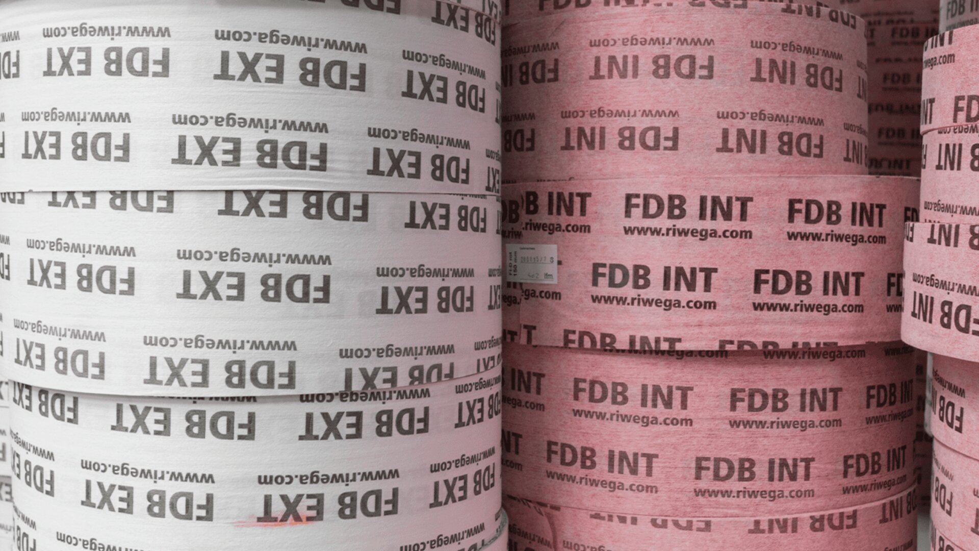 Nahaufnahme gestapelter Materialrollen, mit weißen Rollen auf der linken und rosa Rollen auf der rechten Seite, alle wiederholt mit FDB EXT oder FDB INT und der Website www.riwega.com in schwarzer oder roter Schrift bedruckt.