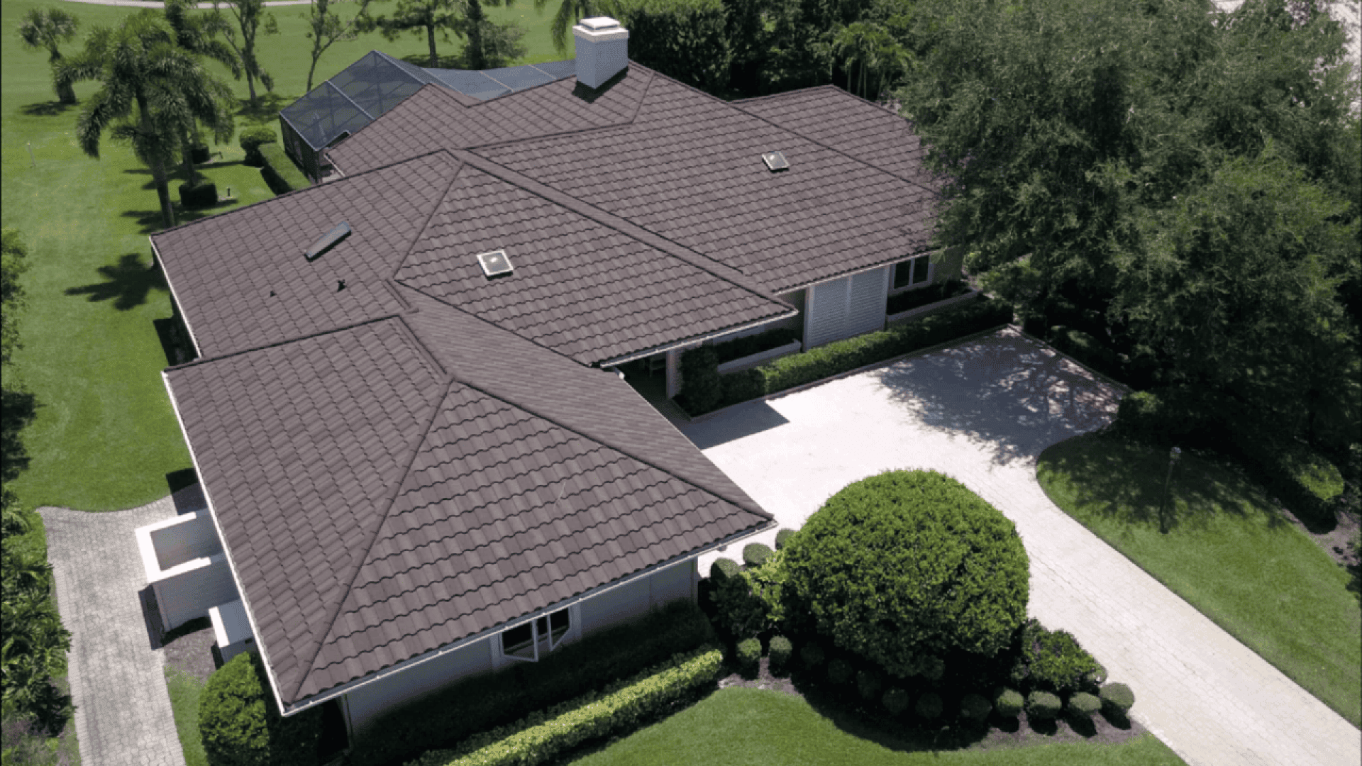 Aerial view of a large, single-story house with a dark, tiled roof, surrounded by neatly trimmed bushes, a driveway, and lush green lawns with trees.