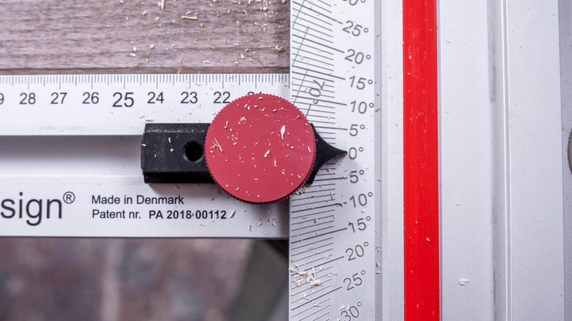 Eine Nahaufnahme eines Tischlermesswerkzeugs, das auf einem Metalllineal mit metrischen Markierungen platziert ist. Bei der 5-cm-Markierung ist eine rote kreisförmige Markierung zu sehen, wobei sich auf dem Werkzeug und der Oberfläche Holzstaub befindet.