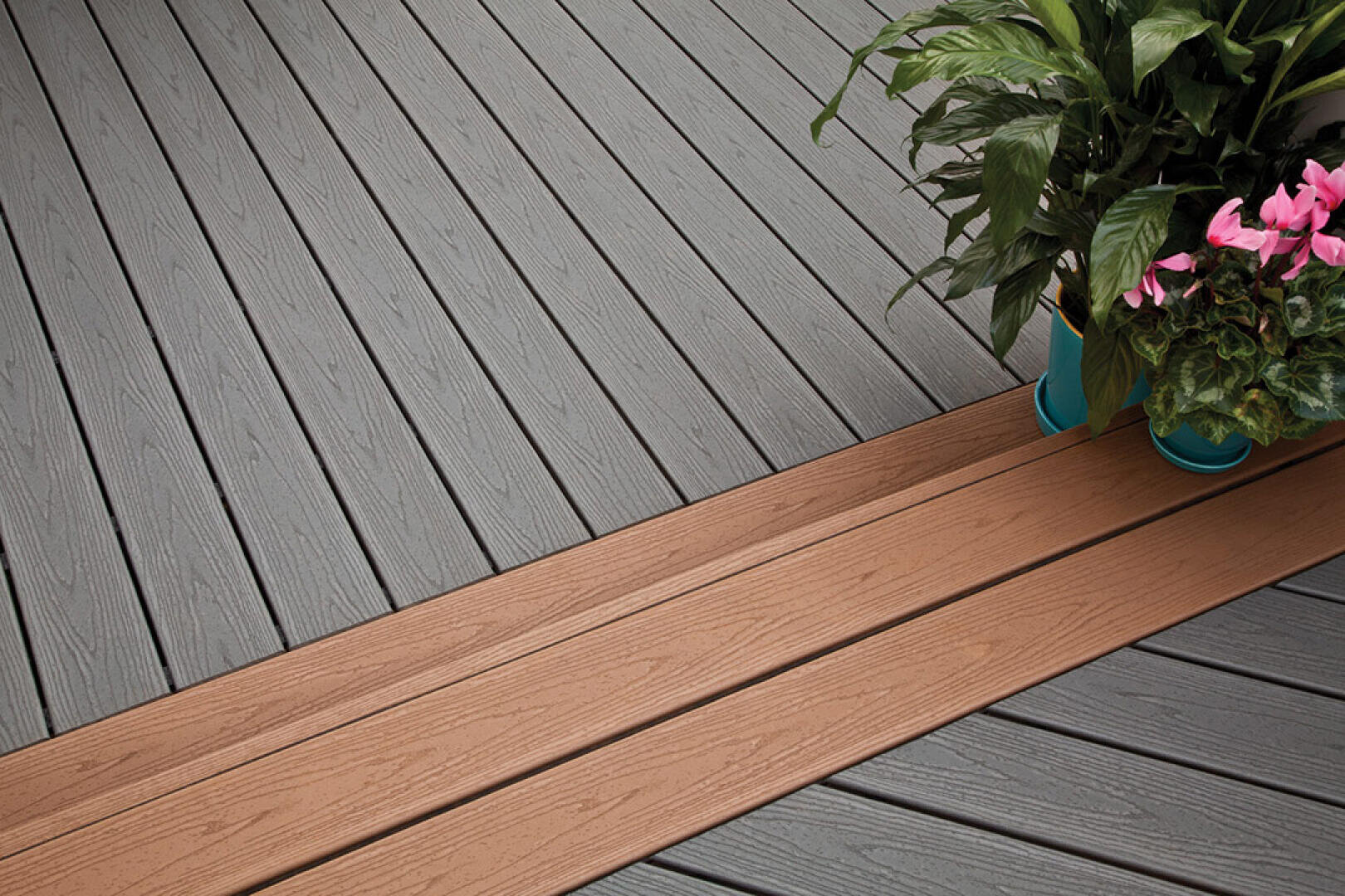Eine Nahaufnahme eines Decks mit diagonal angeordneten grauen und hellbraunen Verbundplatten, mit zwei Topfpflanzen mit grünen Blättern und rosa Blüten auf der rechten Seite.