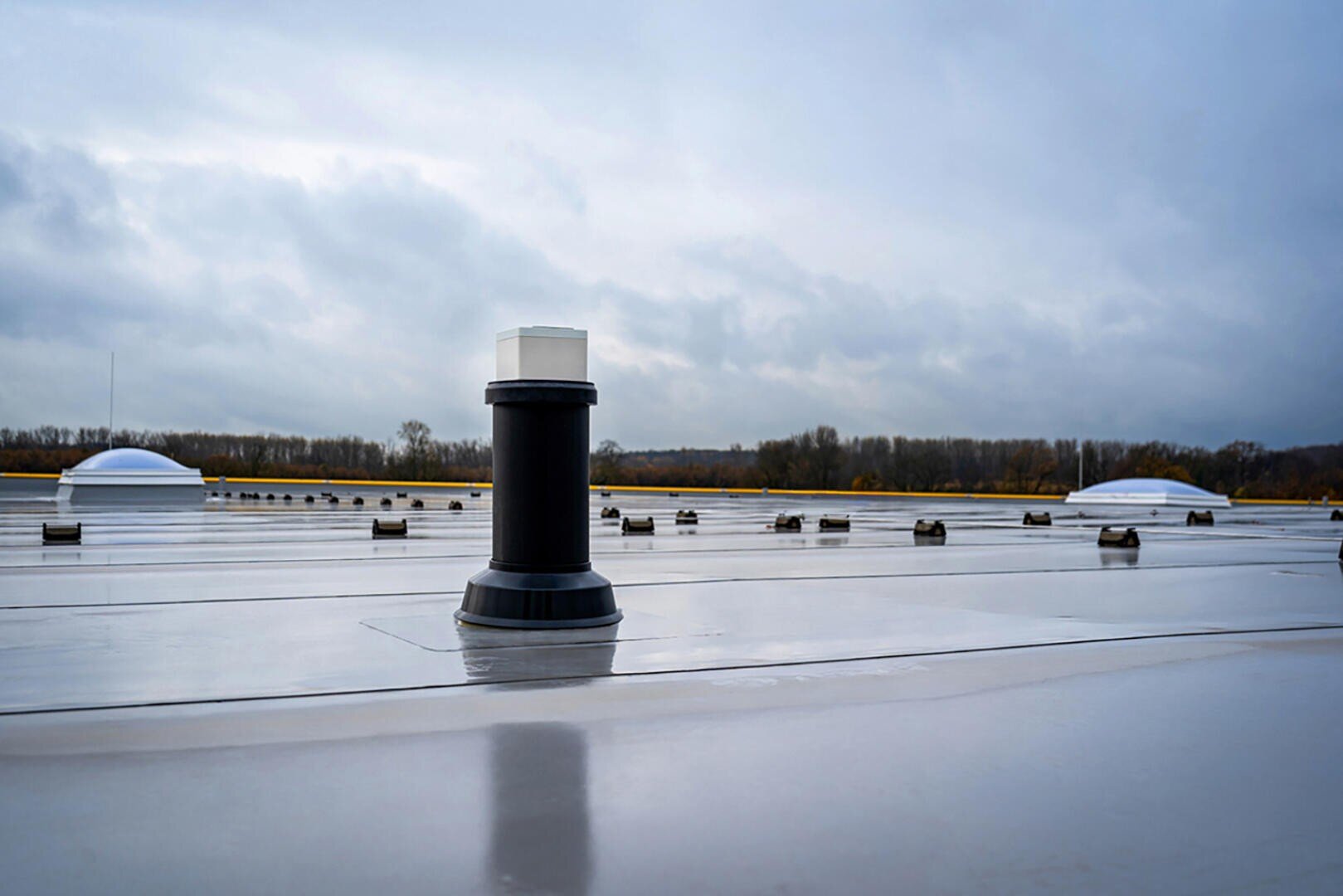 Ein flaches Dach mit mehreren Lüftungsrohren und Oberlichtern, unter einem bewölkten Himmel mit Bäumen im Hintergrund. Die Dachfläche erscheint sauber und reflektierend.