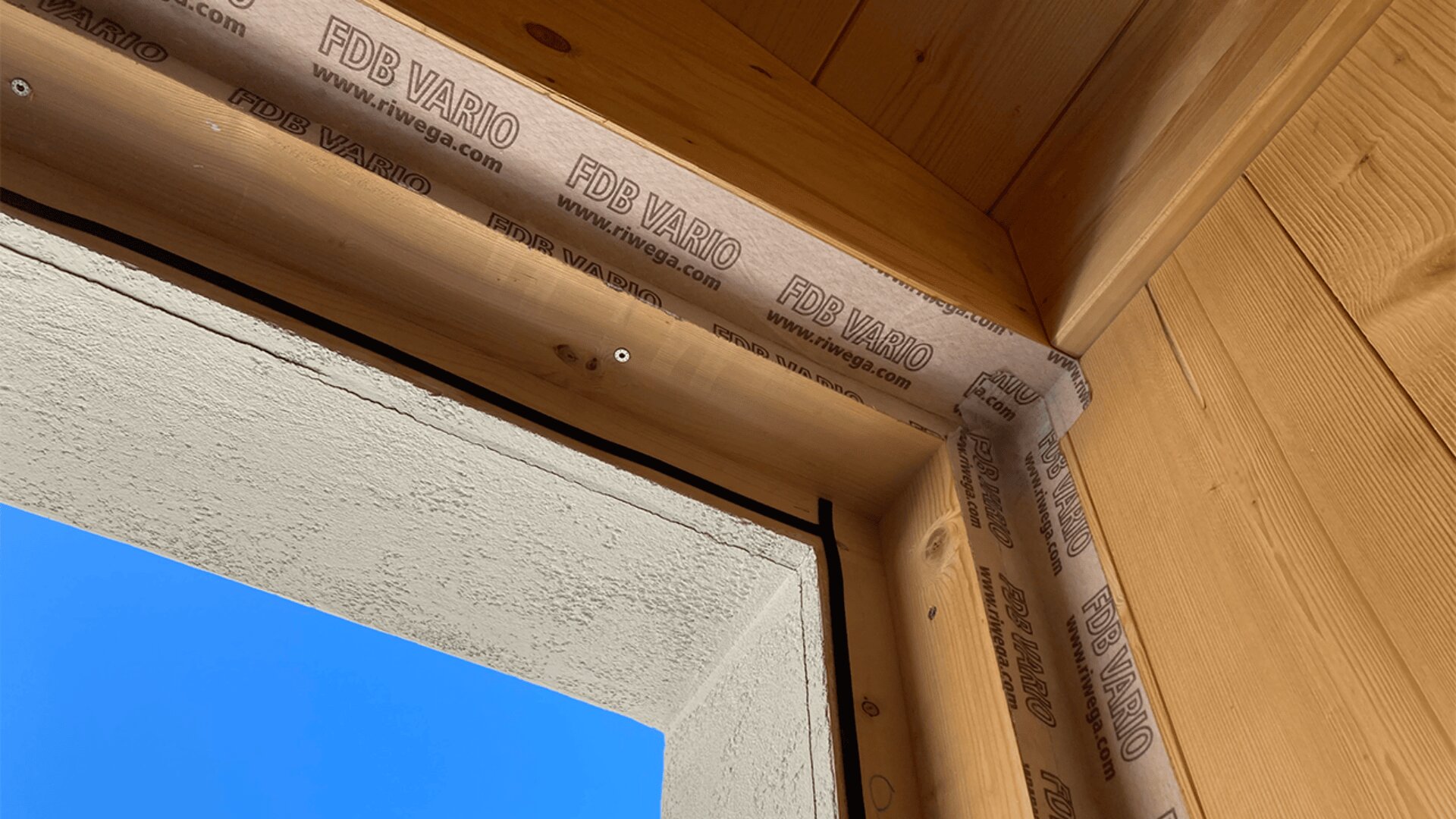 Nahaufnahme eines Holzfensterrahmens mit Isolierband mit der Aufschrift „FDB VARIO“ an den Rändern. Der Rahmen steht vor einer Stuckwand, draußen ist ein strahlend blauer Himmel zu sehen.
