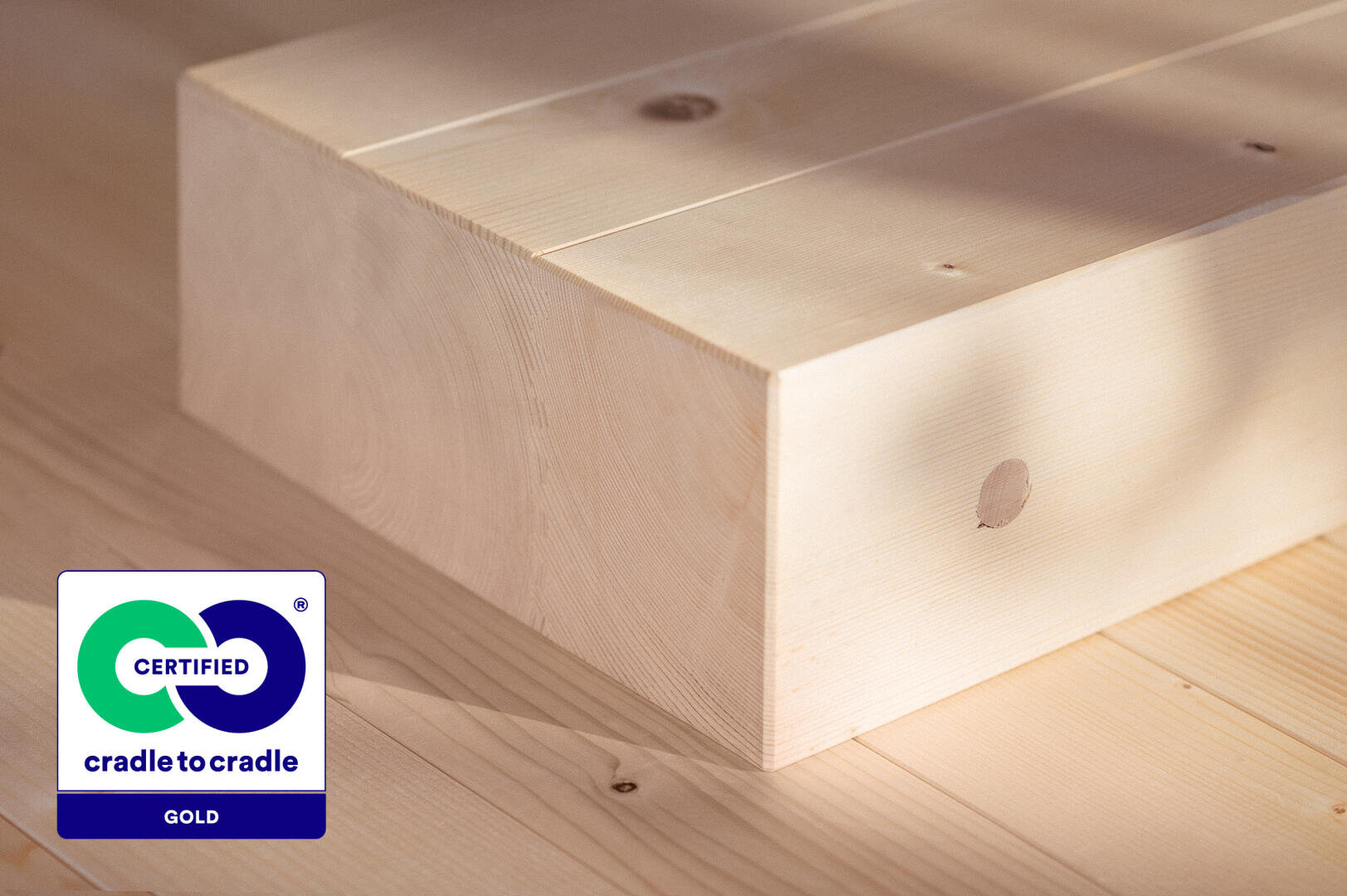 Nahaufnahme heller Holzbalken auf einer Holzoberfläche mit einem Cradle to Cradle Certified Gold-Label in der Ecke, das auf nachhaltige und zertifizierte Materialien hinweist.