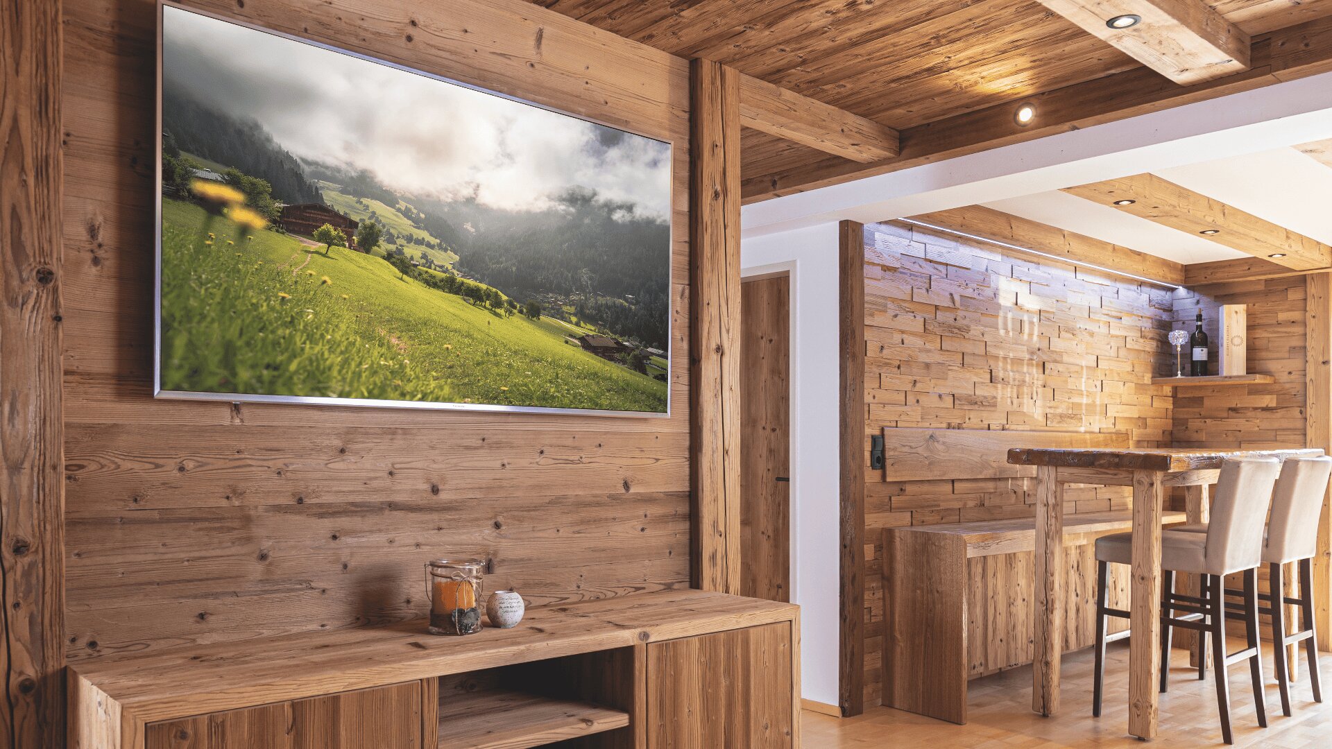 Ein modernes Wohnzimmer mit holzgetäfelten Wänden und Decken, einem Flachbildfernseher mit Berglandschaft, einem hölzernen Esstisch mit Stühlen und einem kleinen Kerzenarrangement auf einem Regal.