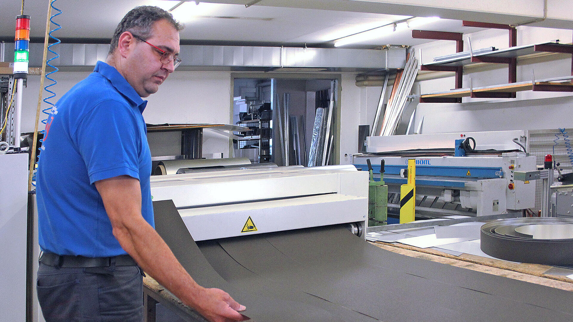 A man in a blue polo shirt operates a machine cutting large sheets of dark material in a factory or workshop setting, surrounded by shelves and industrial equipment.