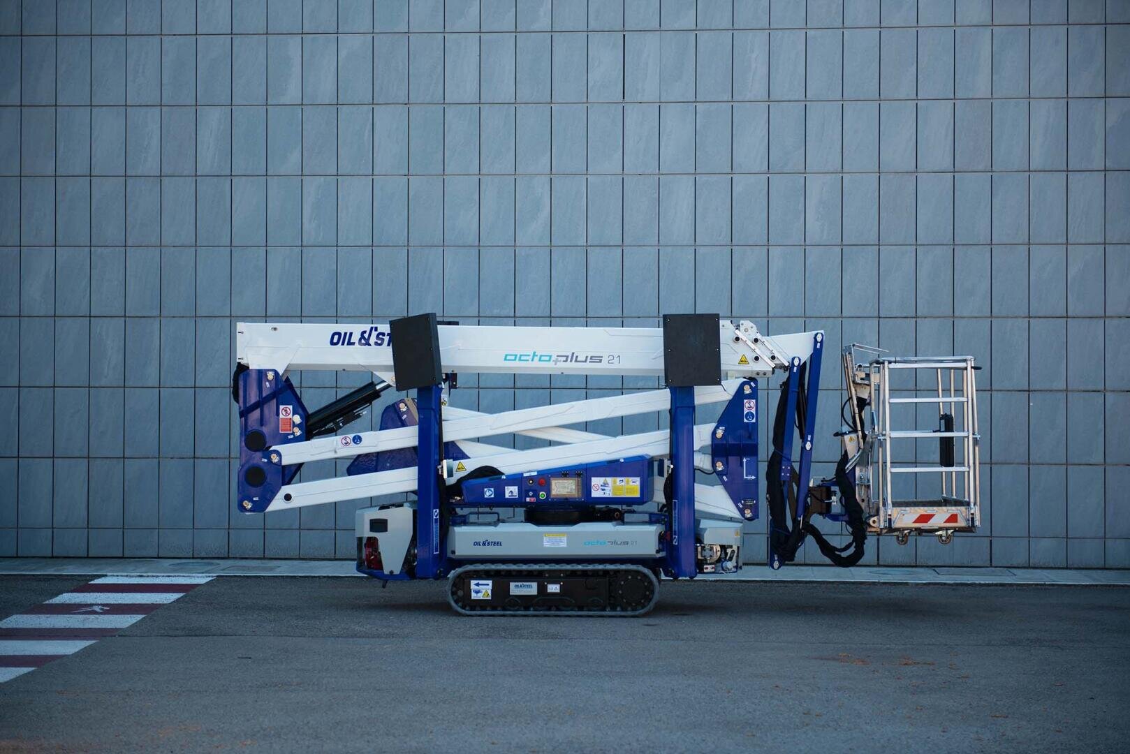 Eine blau-weiße Raupenhebebühne ist vor einer grau gekachelten Wand auf einer gepflasterten Fläche geparkt, mit eingeklapptem Arm und dem Korb auf der rechten Seite.