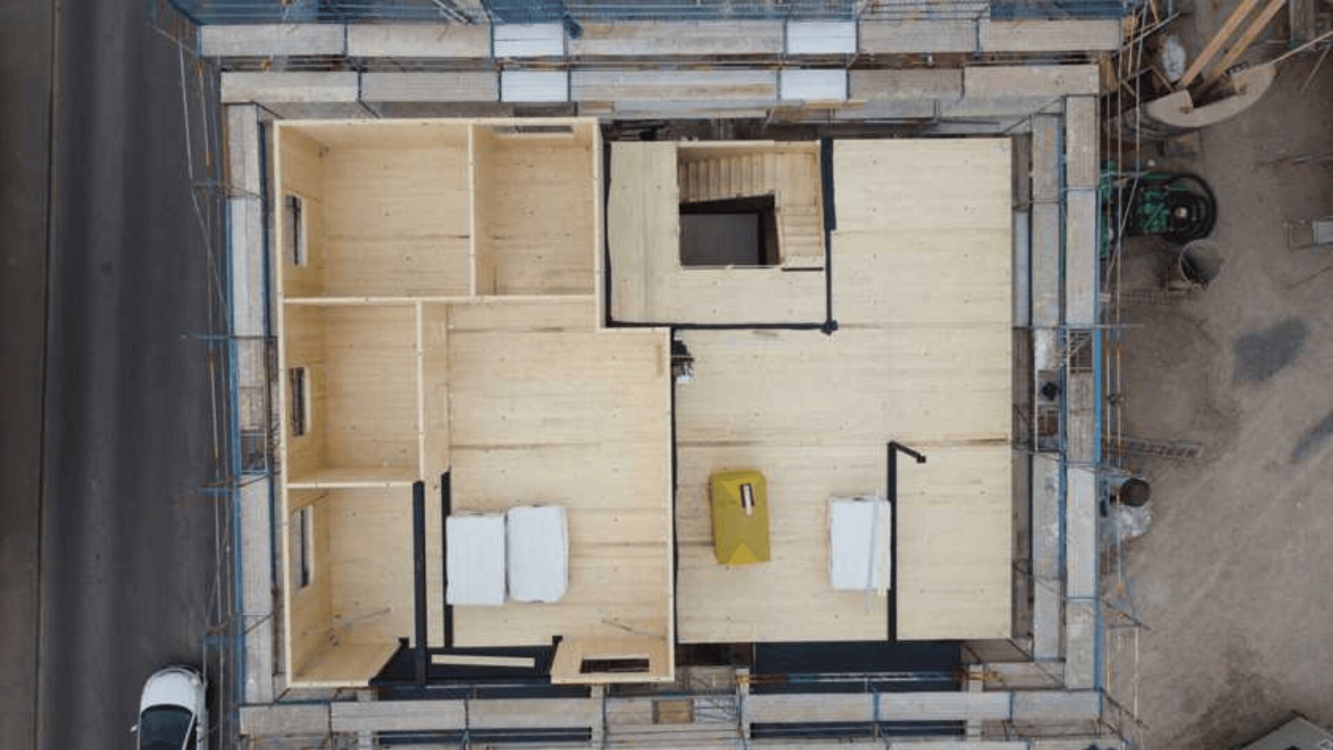 Luftaufnahme eines im Bau befindlichen Hauses. Der Grundriss ist aus Holz, mit mehreren Räumen, teilweisen Innenwänden und einigen Baumaterialien im Inneren der Struktur. Das Gebäude ist von einem Gerüst umgeben.