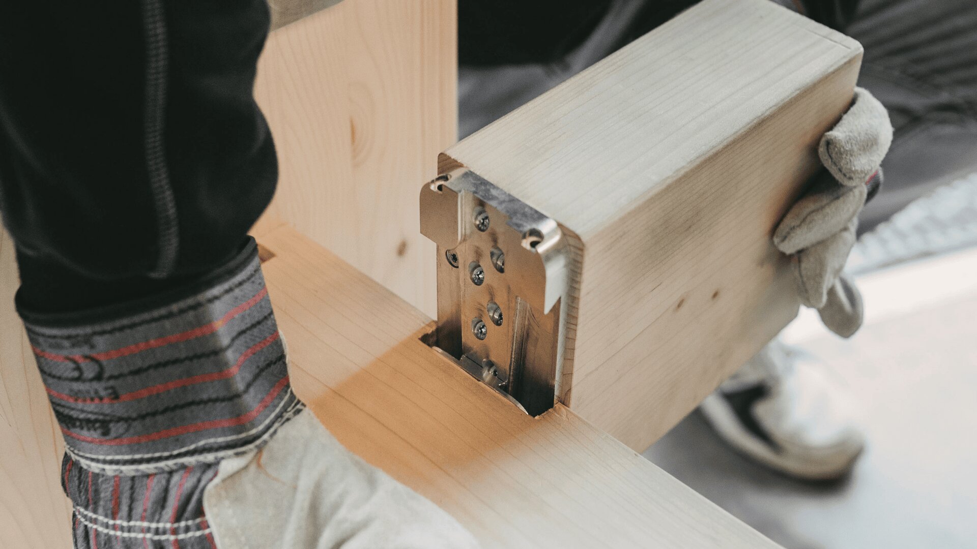 Close-up of a person wearing work gloves assembling wooden furniture, using a metal connector to join two pieces of light-colored wood.