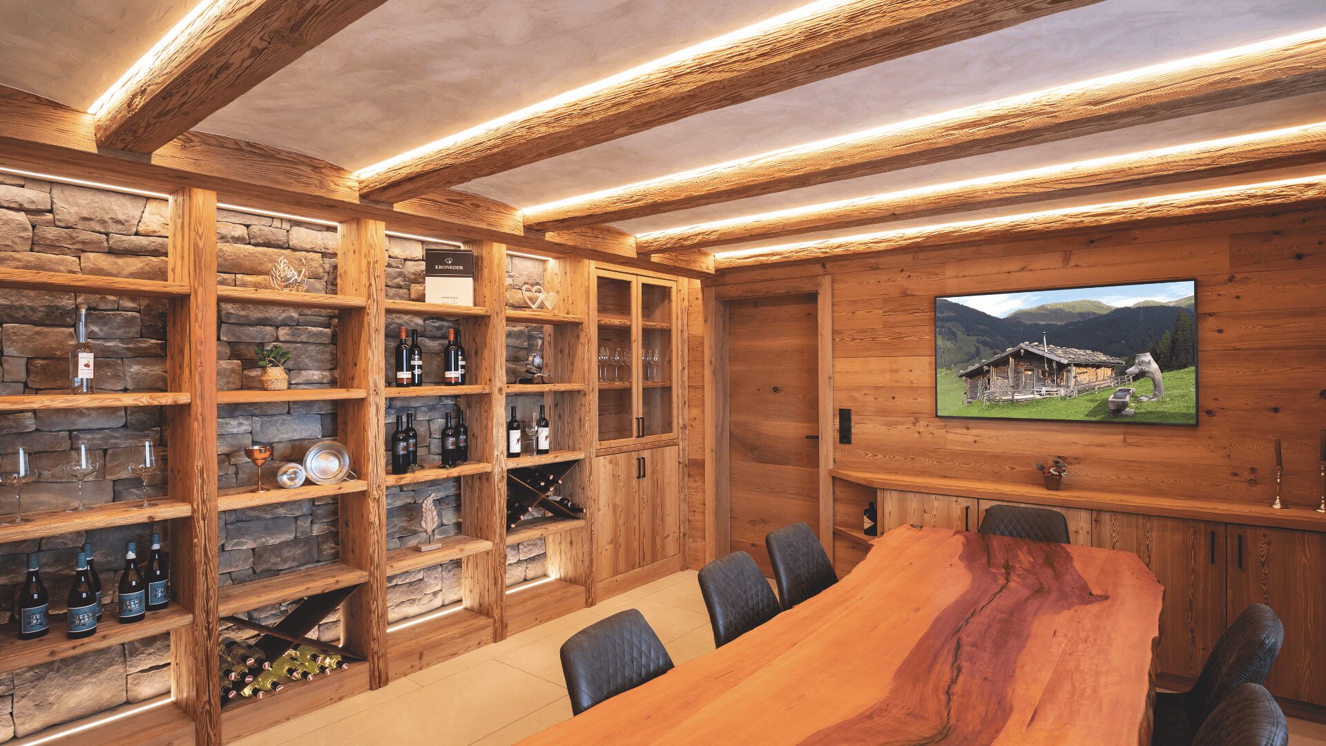 Ein rustikaler Besprechungsraum mit großem Holztisch, schwarzen Stühlen, Holzwänden und -deckenbalken, eingebauten Regalen mit Weinflaschen und einem Fernseher mit einer Berghüttenszene. Warme Beleuchtung unterstreicht die gemütliche Einrichtung.