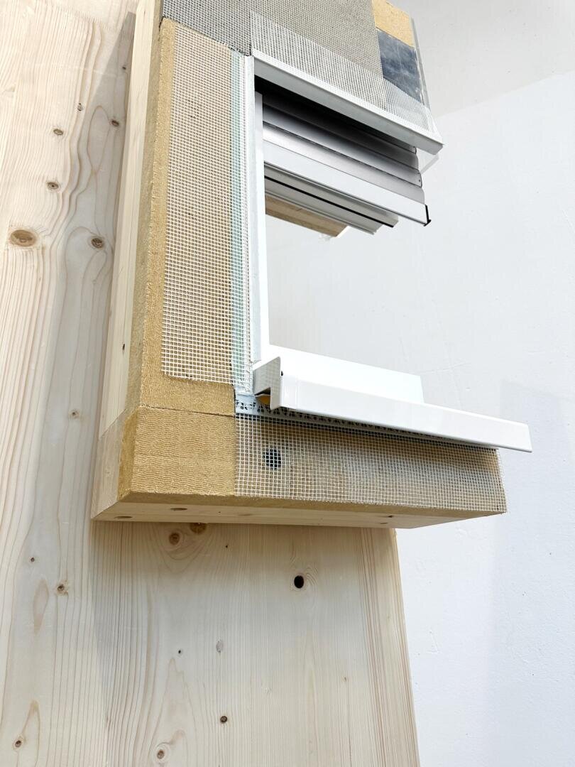 Eine Nahaufnahme eines teilweise konstruierten Fensterrahmens in einer Holzwand mit freiliegender Isolierung, Netzmaterial und weißer Fensterverkleidung. Die Wand- und Fensterelemente sind unbearbeitet und haben ein industrielles Aussehen.