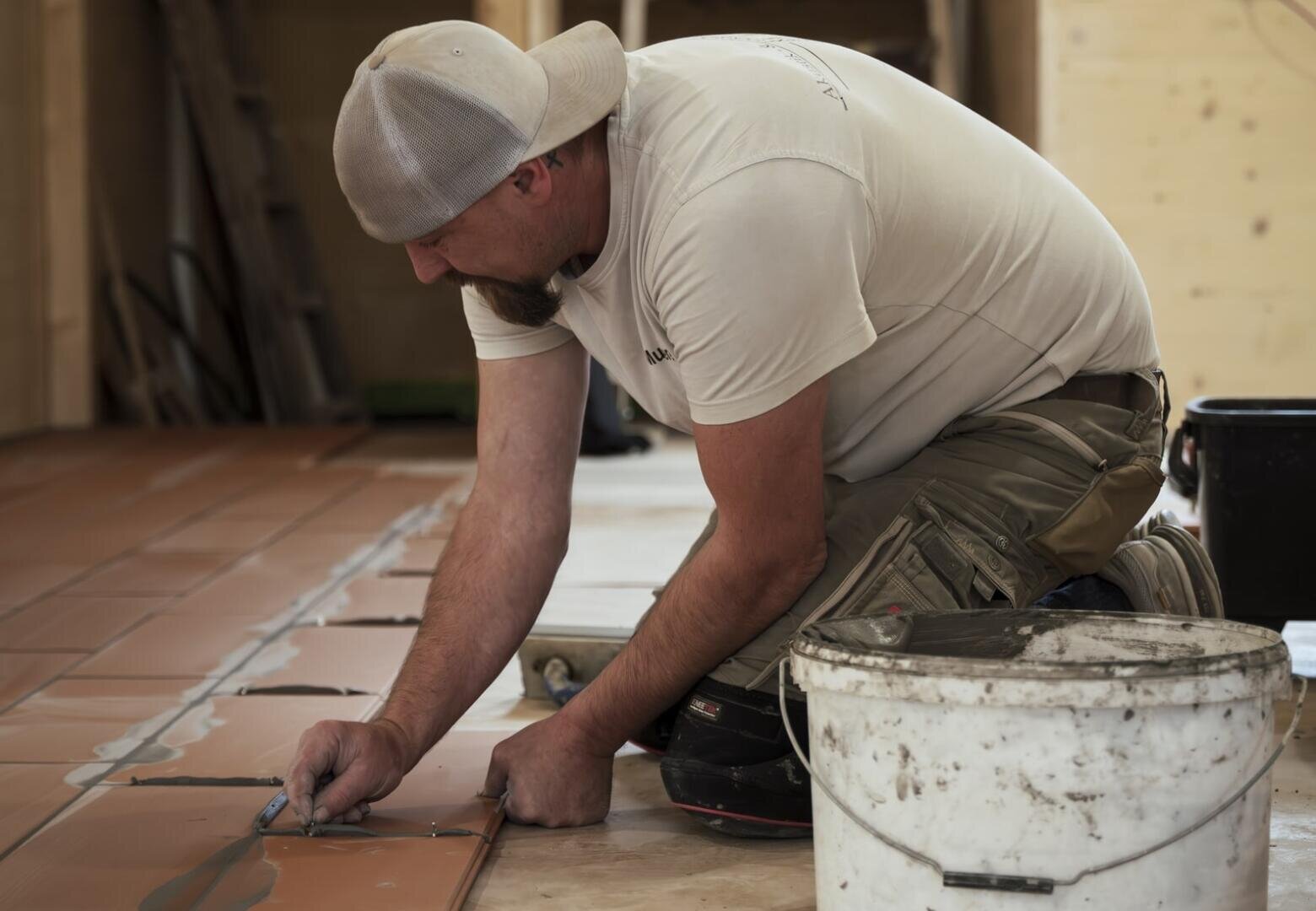 Ein Mann mit einer Schirmmütze und Arbeitskleidung kniet auf dem Boden und verlegt und markiert braune Fliesen. Ein weißer Eimer und Werkzeug stehen neben ihm in einem teilweise fertiggestellten Raum.