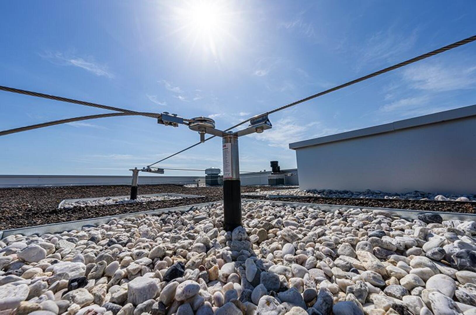 Safety cable system anchored on a gravel-covered flat roof in bright blue sky and sunshine, buildings and roof structures can be seen in the background.