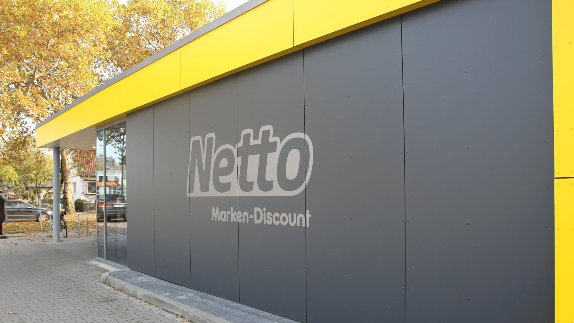 Ein graues Gebäude mit gelber Einfassung zeigt an der Seite das Logo von Netto Marken-Discount. Im Hintergrund sind Bäume mit Herbstlaub und geparkte Autos zu sehen.