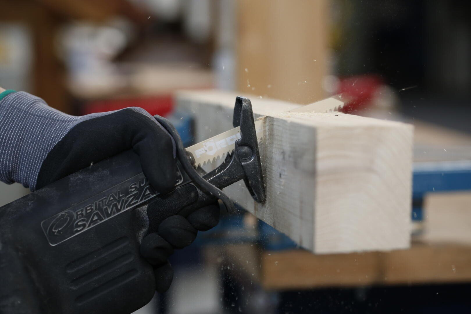 Eine Person mit Handschuhen schneidet mit einer Säbelsäge durch ein Stück helles Holz, wobei Sägemehl um die Schnittstelle herumfliegt.