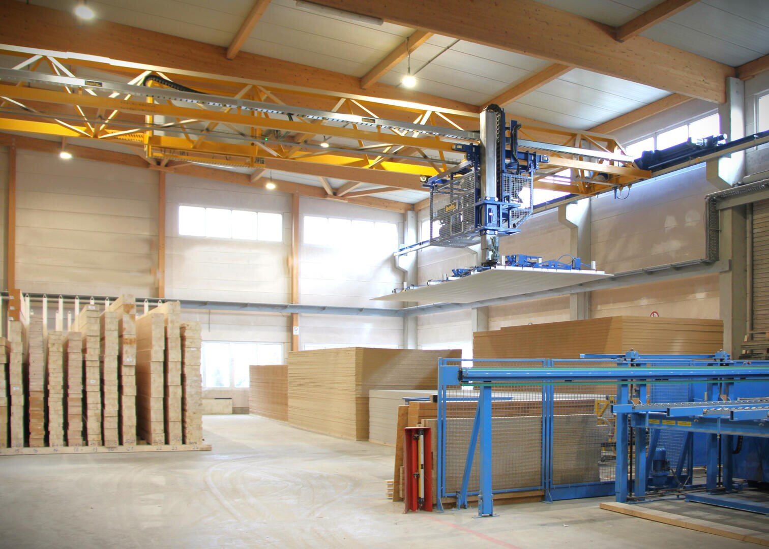 Ein großes, modernes Lagerhaus mit Stapeln von Holzplatten und einem automatischen Brückenkran, der eine Holzplatte anhebt. Der Raum ist hell mit natürlichem Licht aus hohen Fenstern und verfügt über freiliegende Balken und organisierte Materialien.