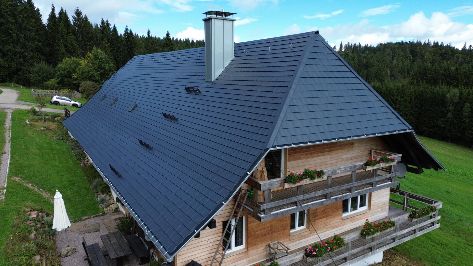 A large wooden house with a steep, dark metal roof sits in a green rural area, surrounded by trees. The balconies have flower boxes, and a white umbrella stands beside picnic tables in the yard.