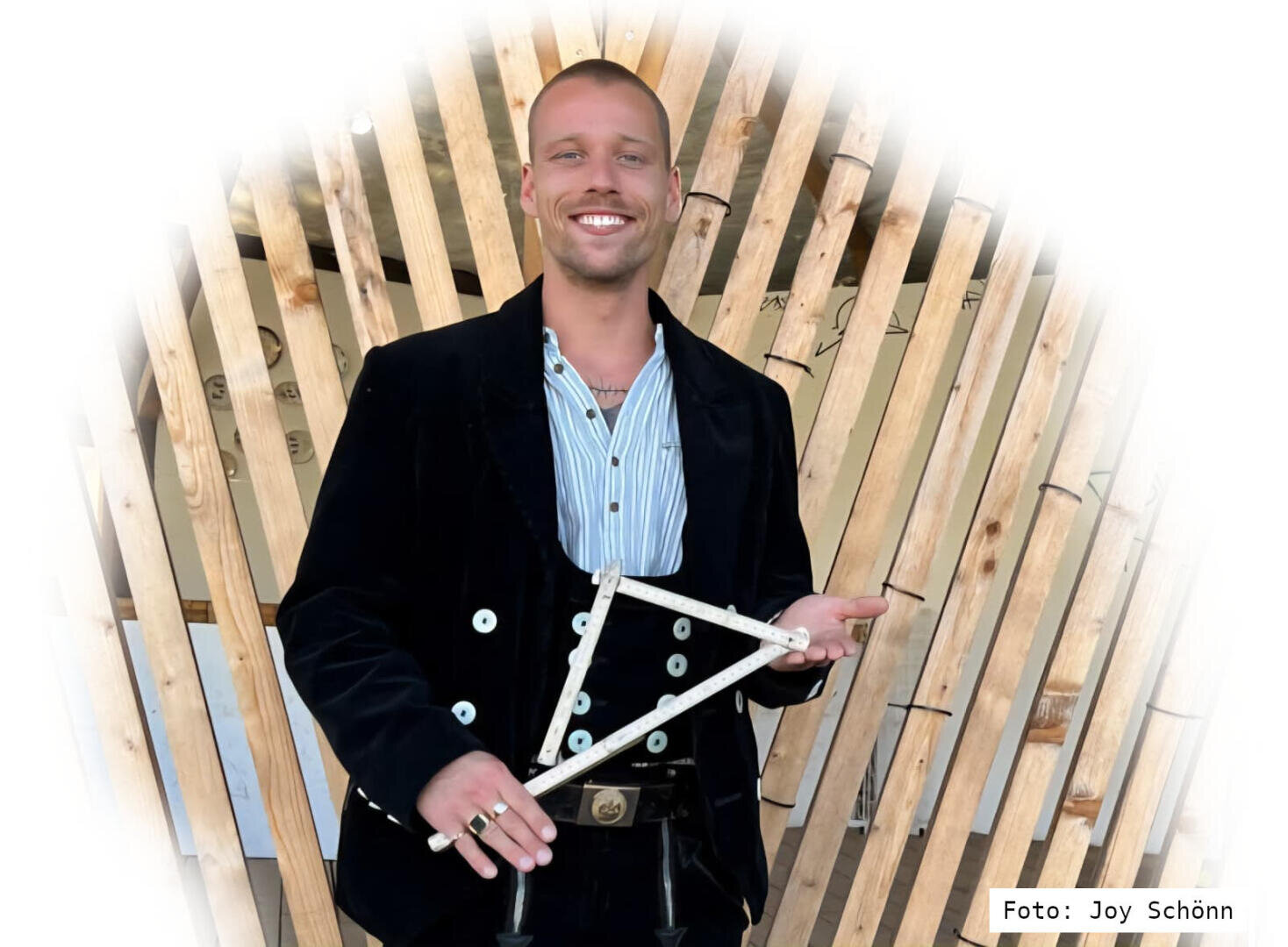 A smiling man in traditional carpenter's clothing holds a folding rule in the shape of a triangle in front of a backdrop of wood and slats. Photo credit: Joy Schönn.