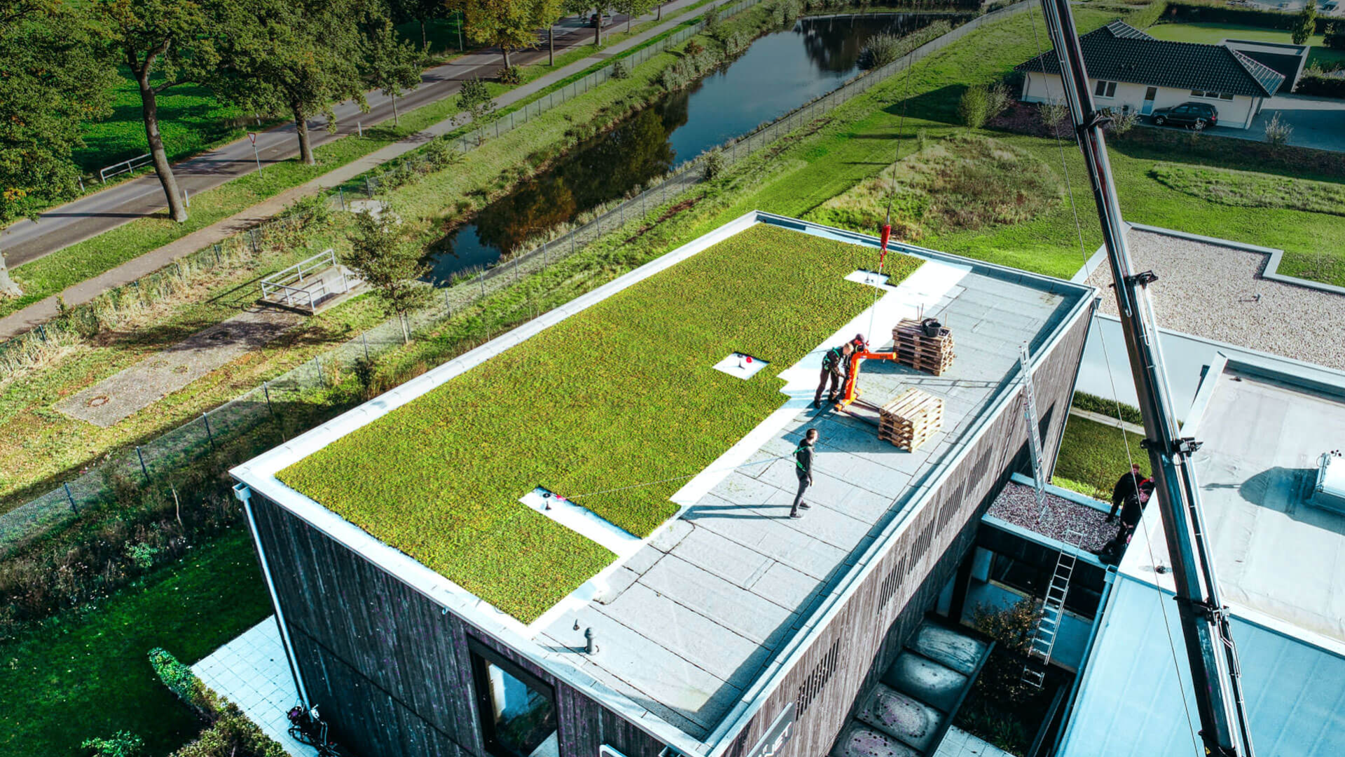 Luftaufnahme von Arbeitern, die ein begrüntes Dach mit Gras auf einem modernen Flachdachgebäude in der Nähe eines Kanals und von Bäumen installieren und einen Kran zum Heben von Materialien verwenden.