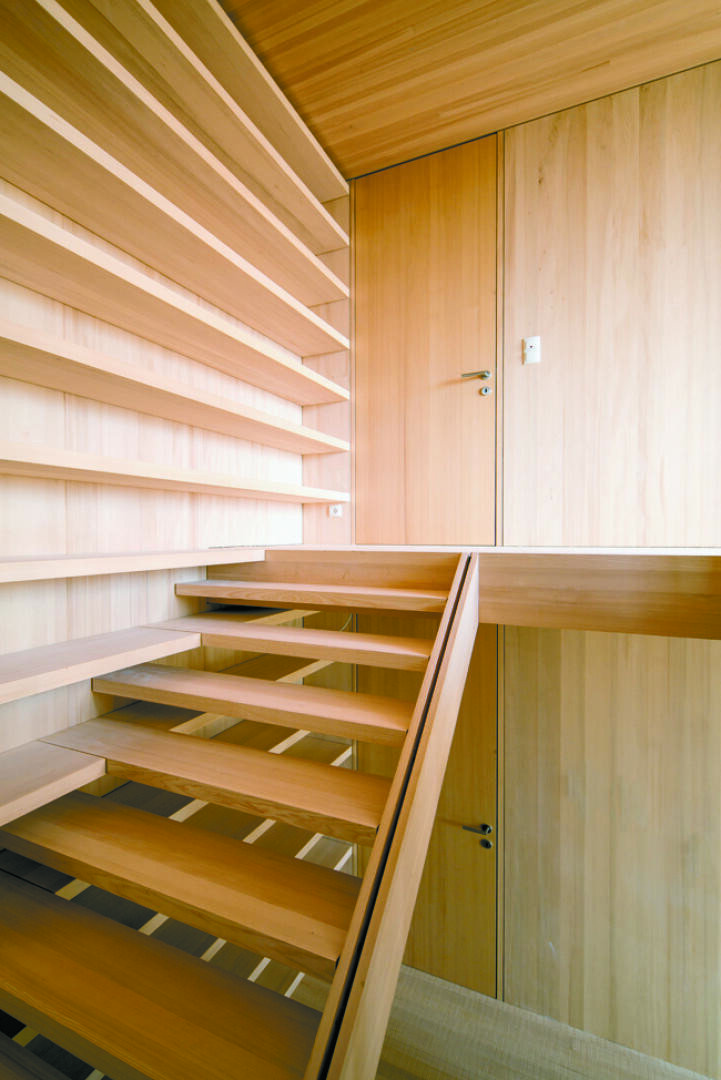 Eine moderne Holztreppe mit offenen Setzstufen, umgeben von glatten, hellen Holzvertäfelungen an Wänden und Decke, führt zu einer geschlossenen Holztür im Obergeschoss. Der Raum wirkt minimalistisch und hell.