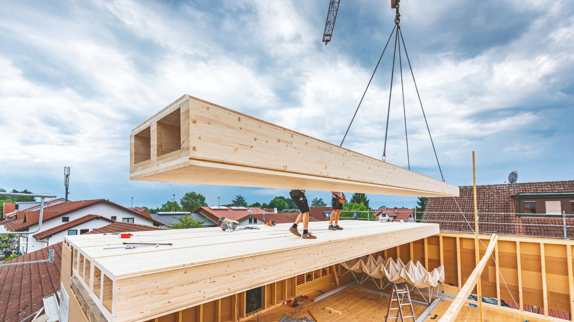 Ein großer Holzbalken wird von einem Kran auf das Gerüst eines im Bau befindlichen Gebäudes gehoben. Zwei Arbeiter führen ihn. Im Hintergrund sind Häuser und ein bewölkter Himmel zu sehen.