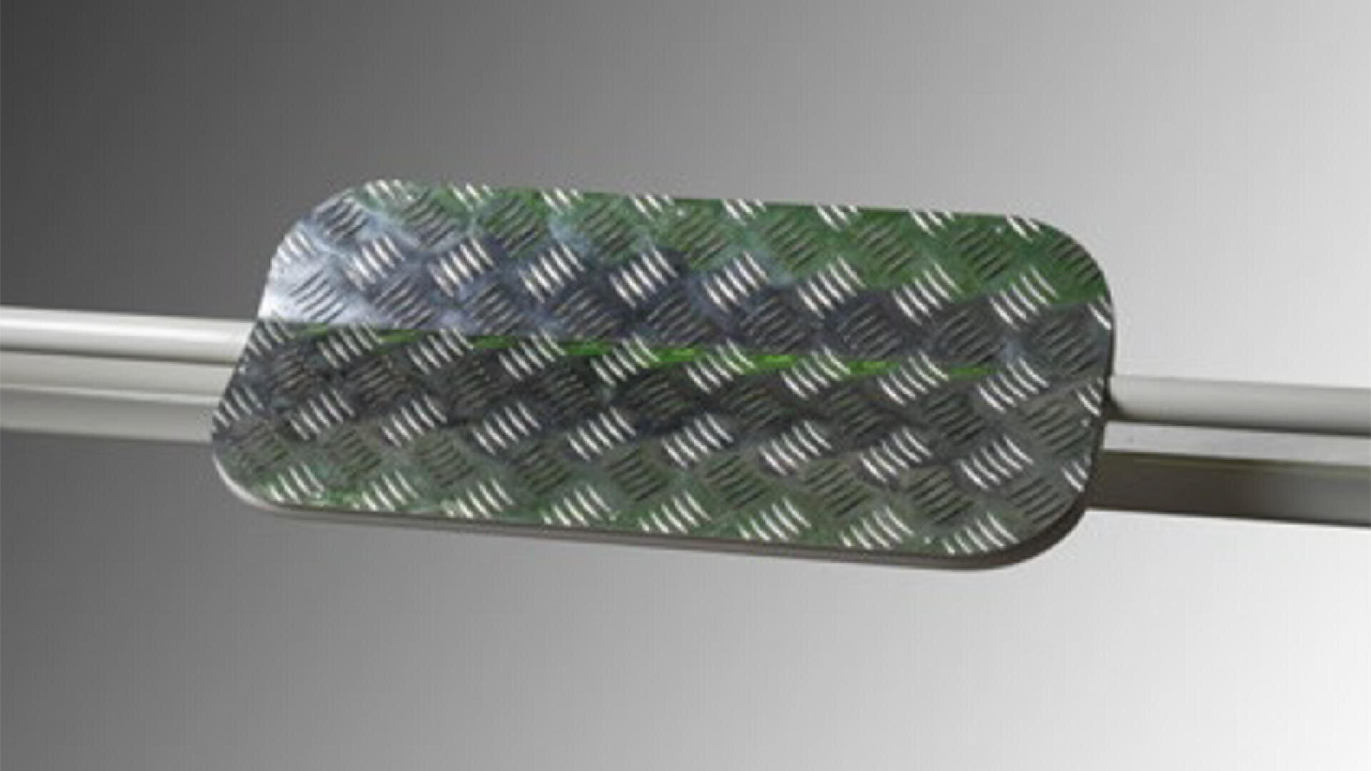 Eine rechteckige Metallplatte mit abgerundeten Ecken und einem erhabenen Rautenmuster, montiert auf parallelen Metallstäben vor einem grauen Hintergrund mit Farbverlauf.