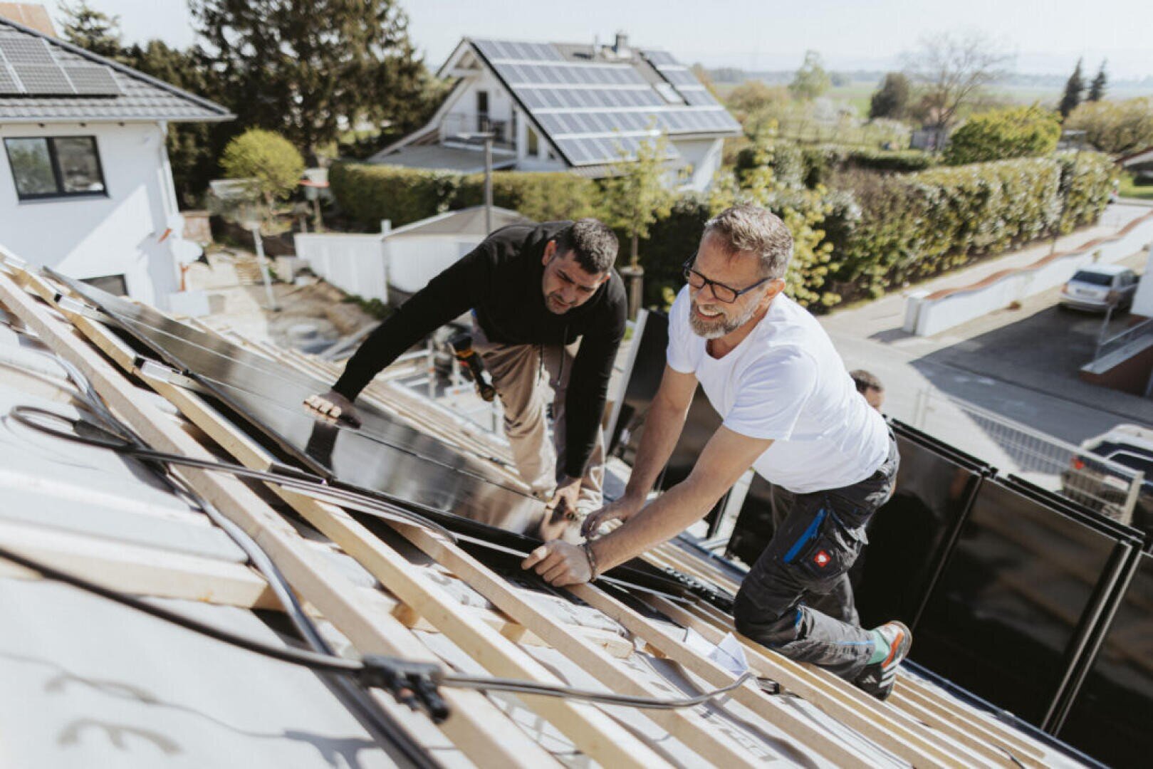 Zwei Männer installieren an einem sonnigen Tag Sonnenkollektoren auf einem schrägen Hausdach in einer Wohngegend. Sie arbeiten mit Werkzeugen und scheinen sich auf ihre Arbeit zu konzentrieren, während auf den Dächern in der Nähe Sonnenkollektoren zu sehen sind.
