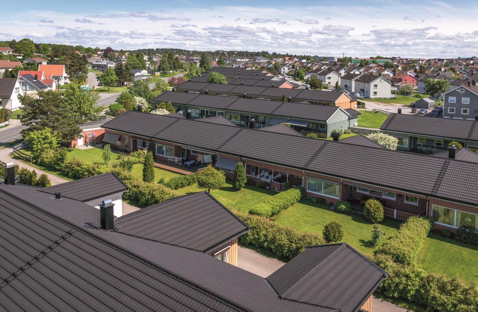 Eine Vorstadtsiedlung mit Reihen einstöckiger Häuser mit schwarzen Ziegeldächern, grünen Rasenflächen, Sträuchern und Bäumen. Der Himmel ist teilweise bewölkt und viele Häuser sind in der Ferne zu sehen.