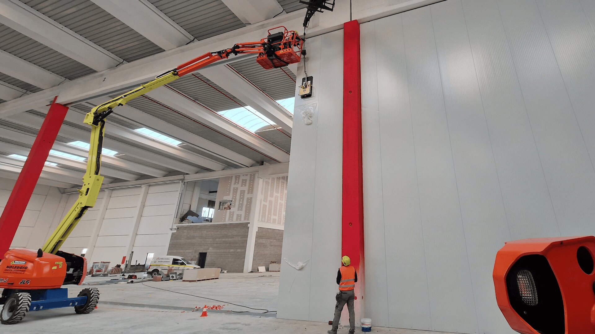 A worker in a safety vest and helmet stands beside a tall, red beam inside a large industrial building, while another worker operates a yellow and orange boom lift to access the top of the wall.